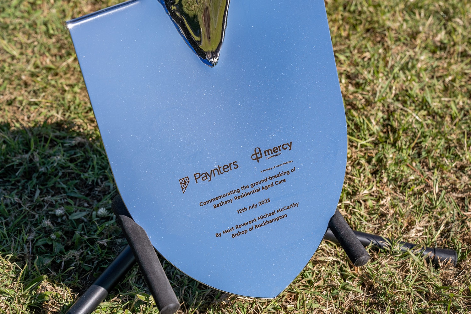 A close-up of the shovel used for the groundbreaking. It is a shiny metal with the Paynters and Mercy Community logo. Under the logo reads "Commemorating the ground-breaking of Bethany Residential Aged Care. 12th July 2023. By Most Reverent Michael McCarthy Bishop of Rockhampton.