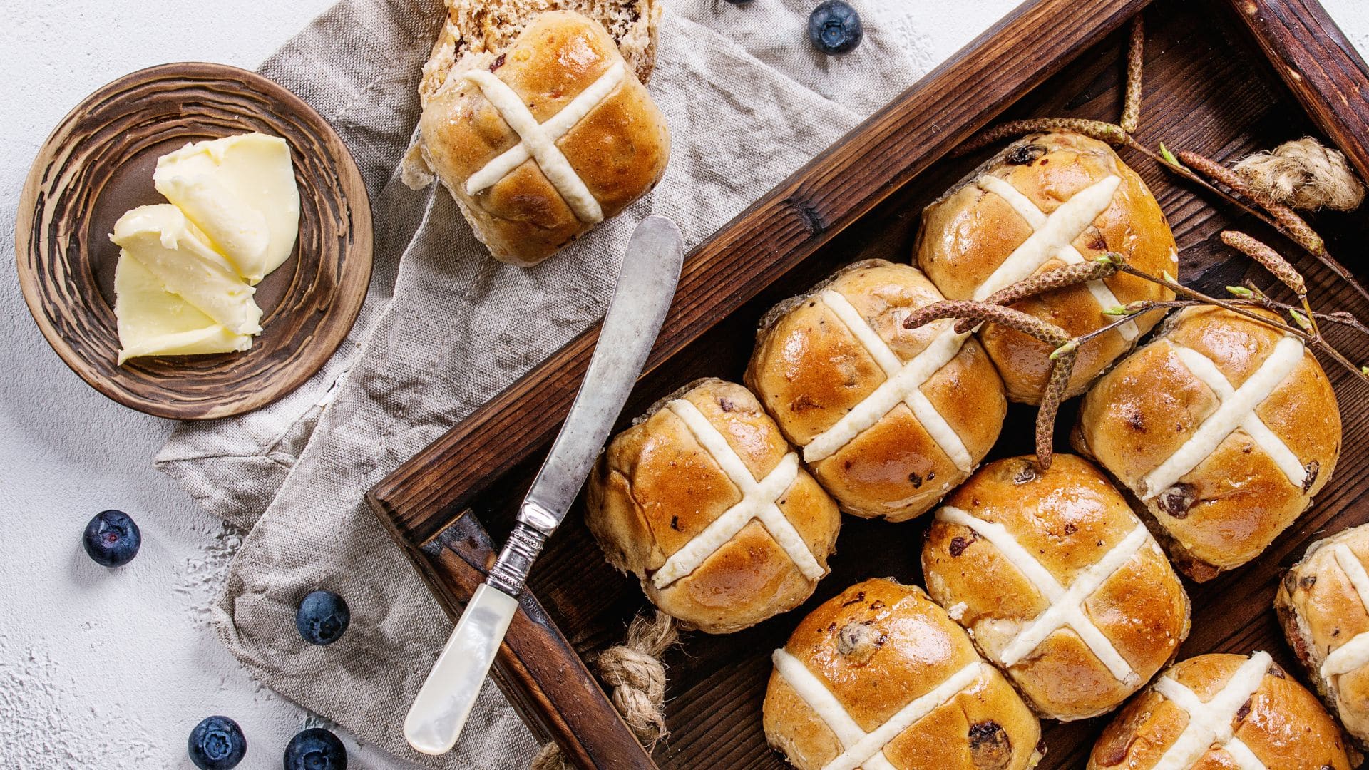 A batch of freshly baked hot cross buns sit on a table with a plate of butter and blueberries.