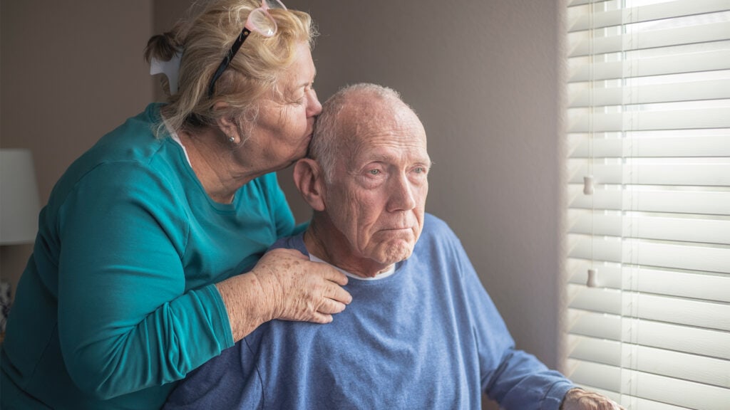 A daughter rests her hands on her elderly fathers shoulders while kissing him affectionately on the head. He's looking somewhere off camera with a sombre expression.