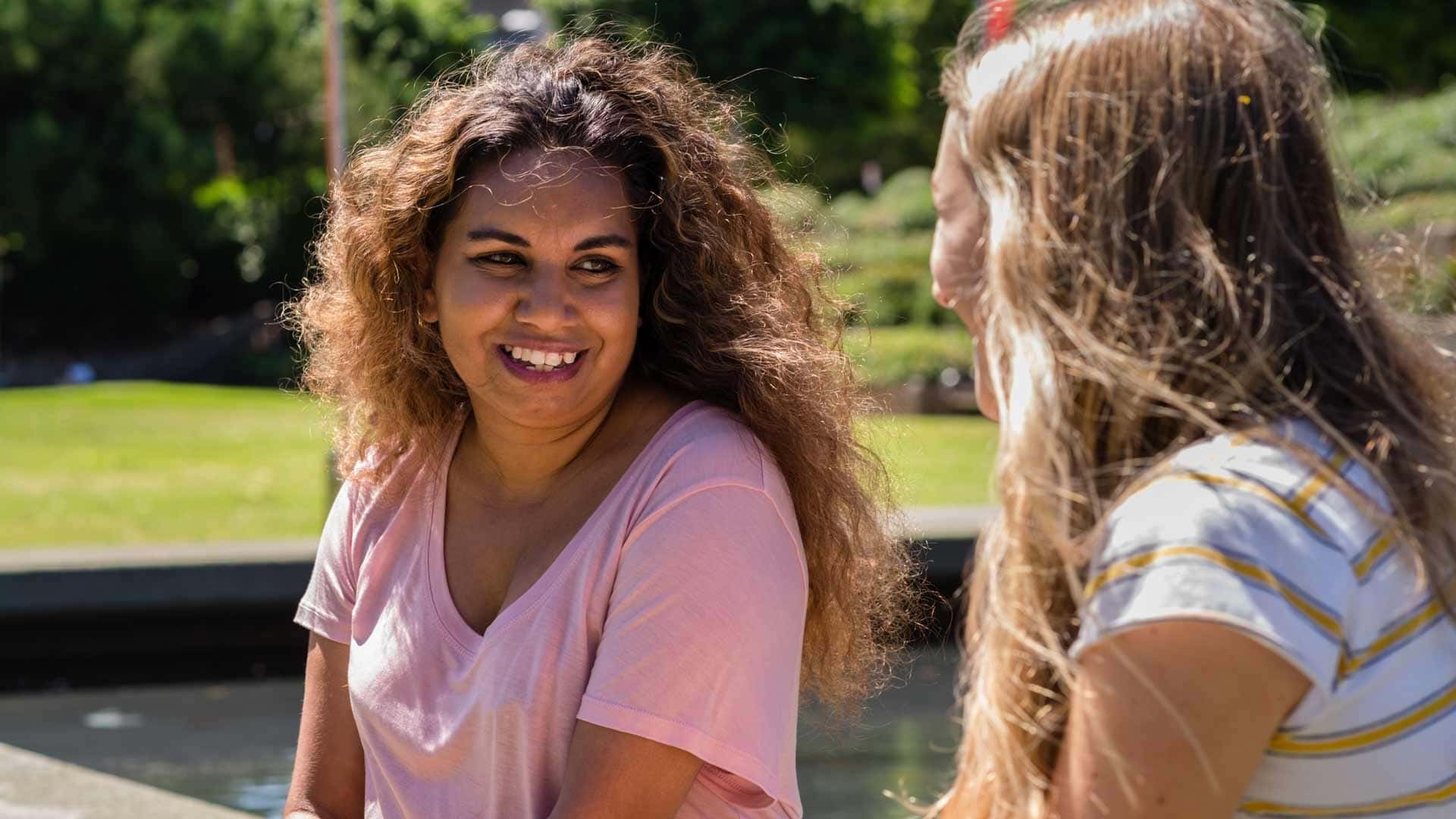 First Nations woman smiles and chats to a blonde woman while sitting in a park.