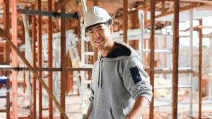 A young man wearing a hard hat and grey hoodie smiles at the camera. He's standing in a construction site which appears to be for a house or building.