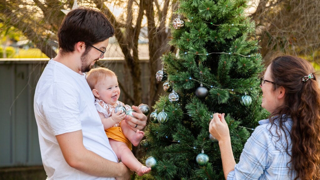 A mother and father decorate a Christmas tree in the backyard with their baby. The father is holding the baby who is holding an ornament and smiling at their mother.