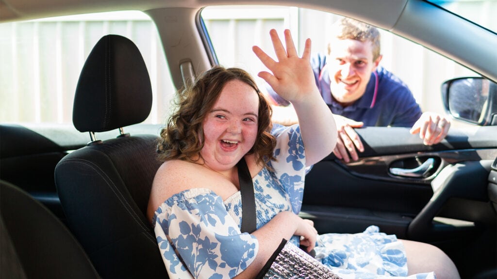 A young woman sits in the drivers seat smiling and waving to some one off camera. A young man who is her support worker leans on the open car window behind her, smiling at her.