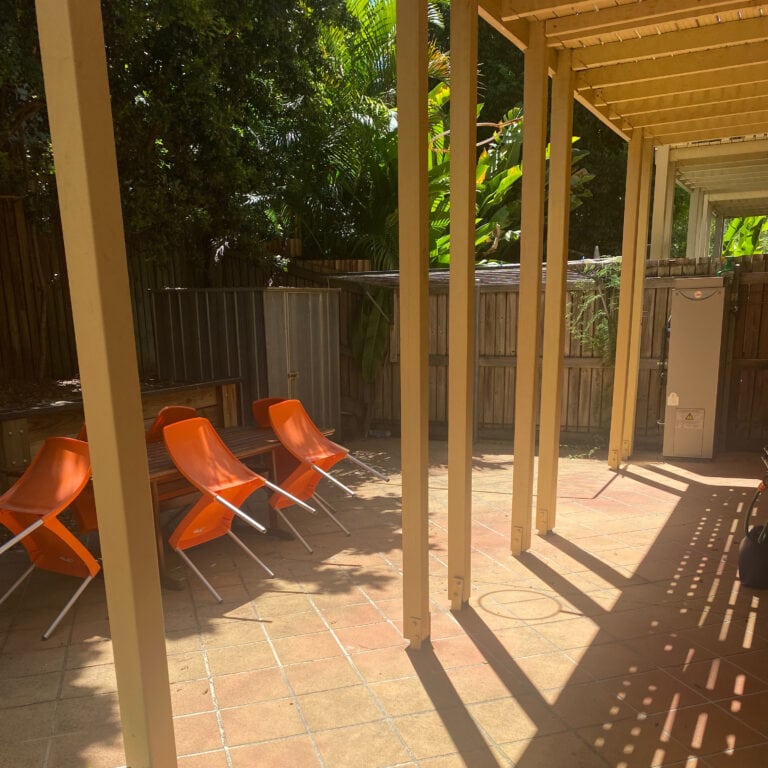 A sunny back patio paved with terracotta tiles is shown. The are is surrounded by large palms and a large wooden fence.