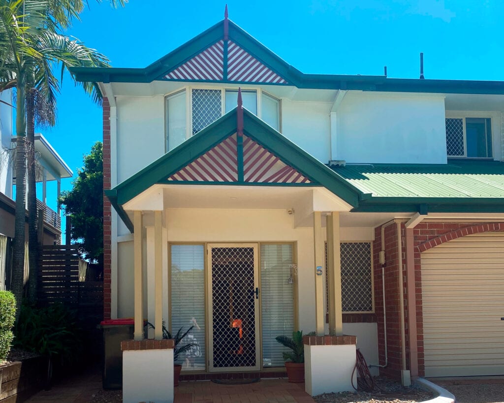 The front of the SIL home in Corinda. The front entrance and a second floor window can be seen. The home exterior is yellow with dark green and red trimmings and accents.