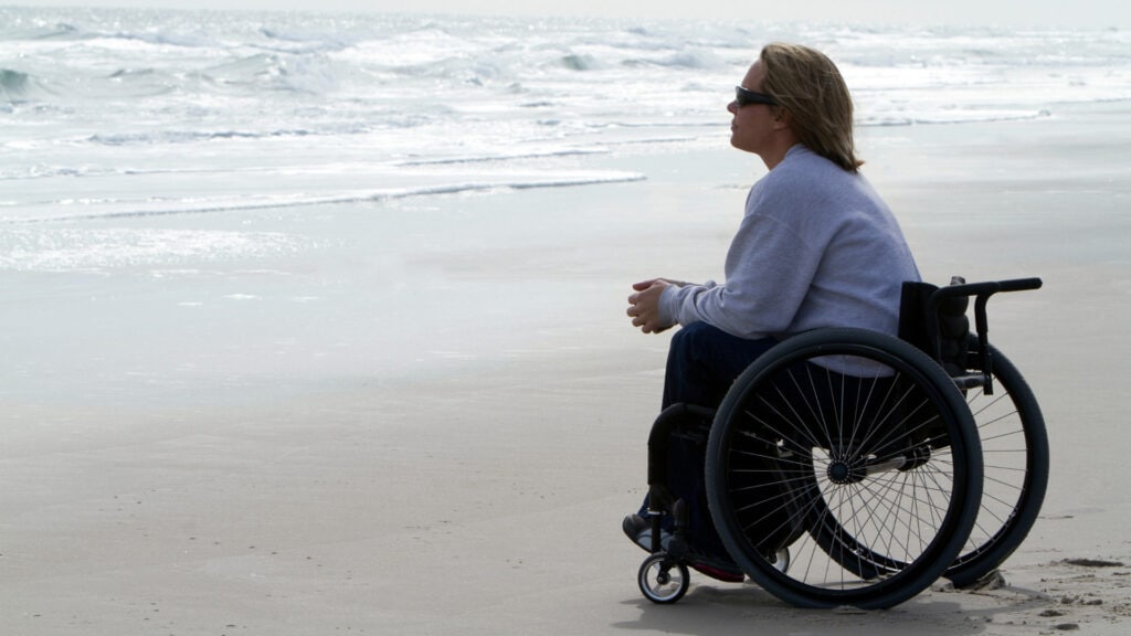 A woman sitting on a beach in a wheelchair looks out over the water.