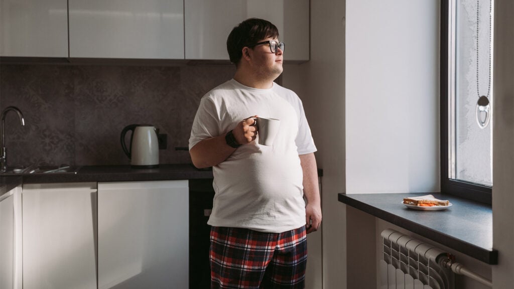 A young man with down syndrome looks out the kitchen window while having his morning coffee.