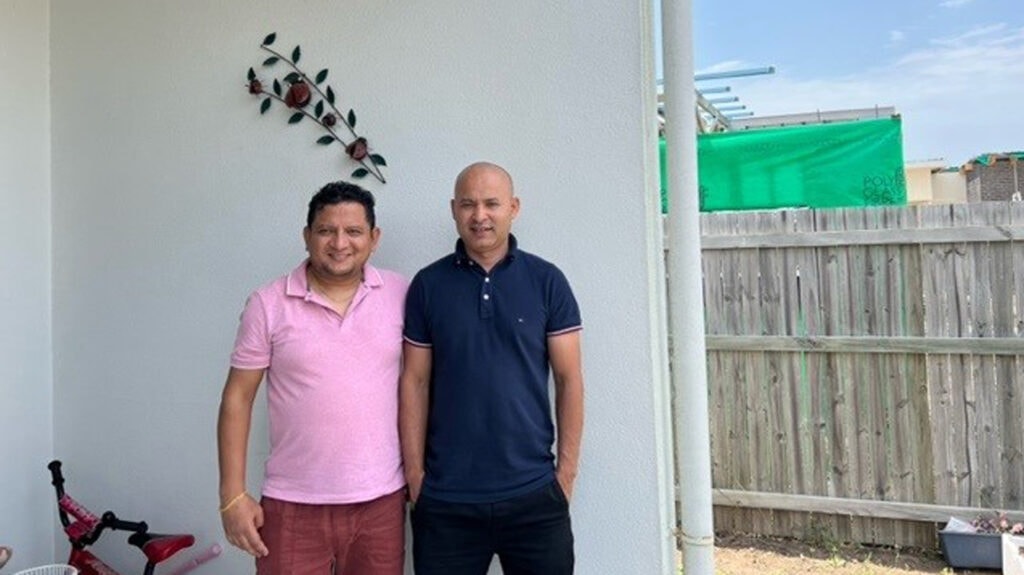 Dinesh and Saroj stand together smiling at the camera. They're standing against a white wall in a backyard garden,