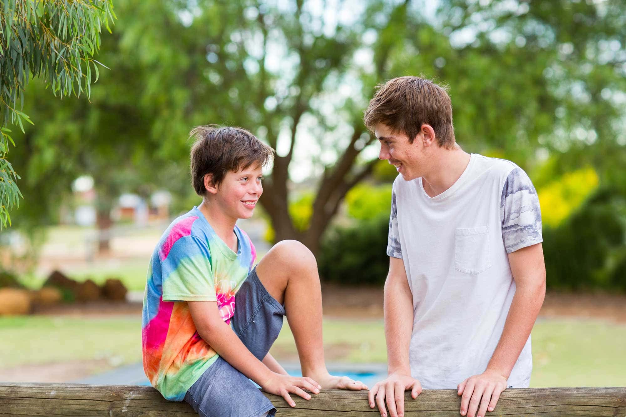 A young boy is sitting on a fence talking with his older brother who is leaning on the fence. They're both smiling.