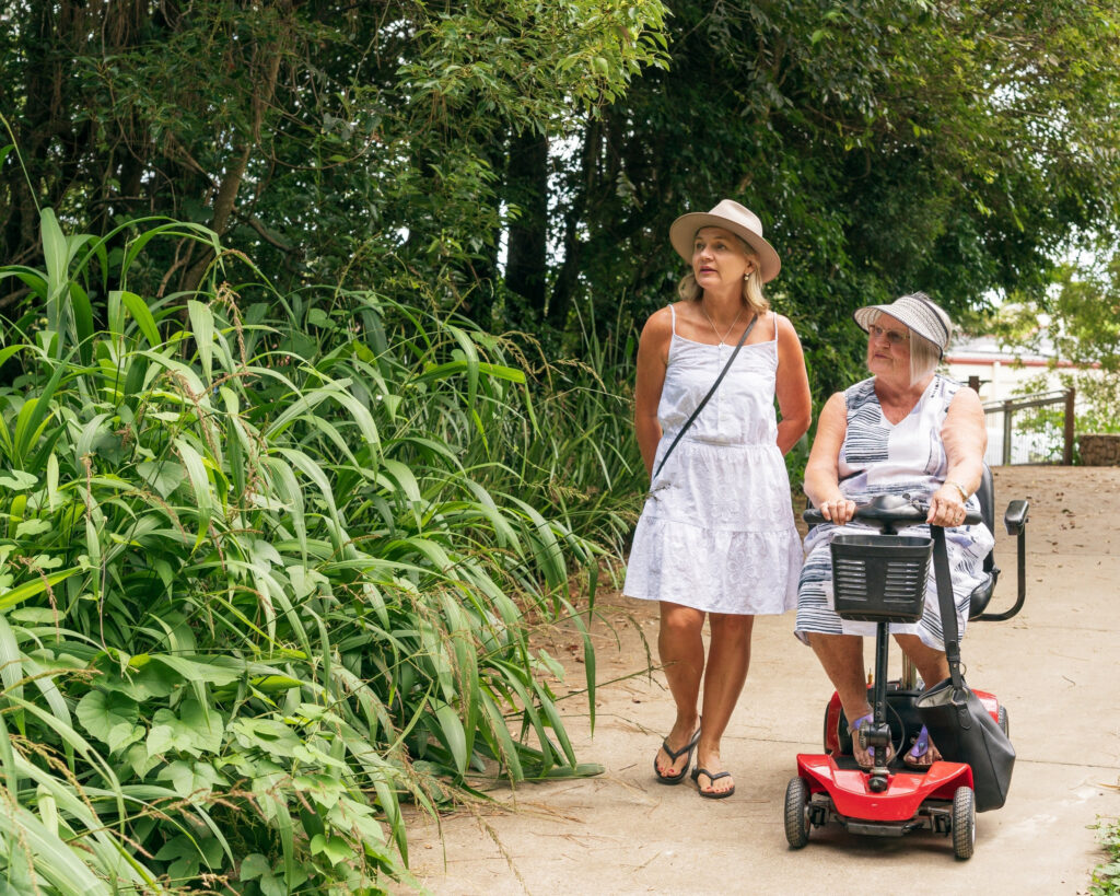 Two women, one on a mobility scooter, chatting and walking along garden pathway
