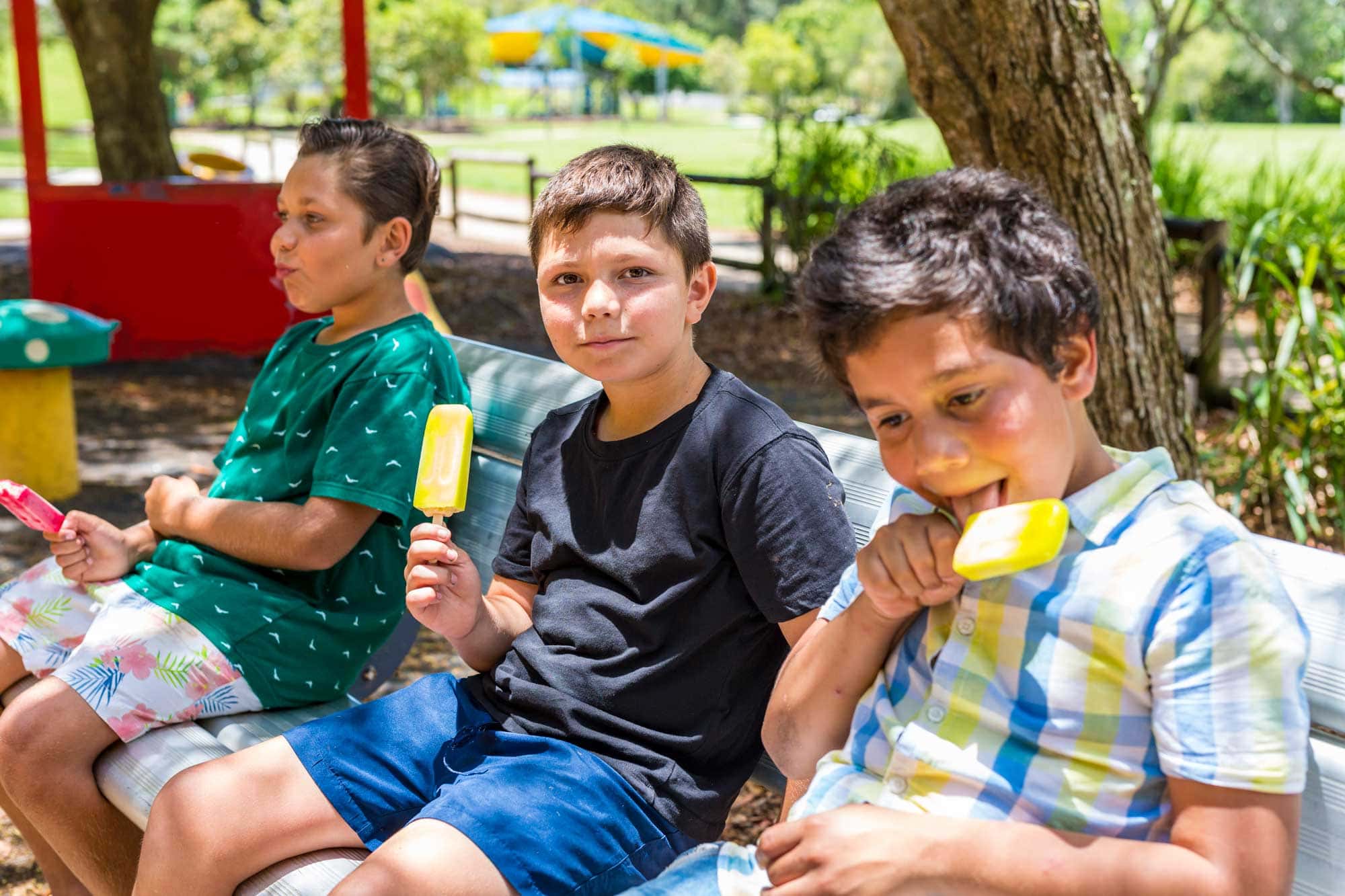 A young boy with an ice block smiles at the camera. He's sitting on a park bench with his two brothers who are looking away from the camera eating their own ice blocks.