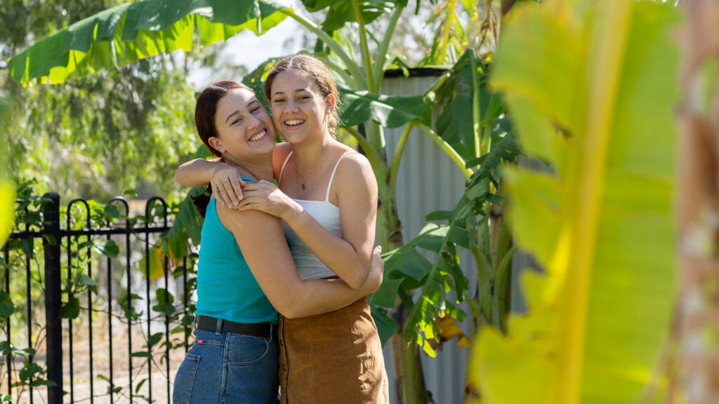 Two sisters smile at the camera in their backyard while hugging.