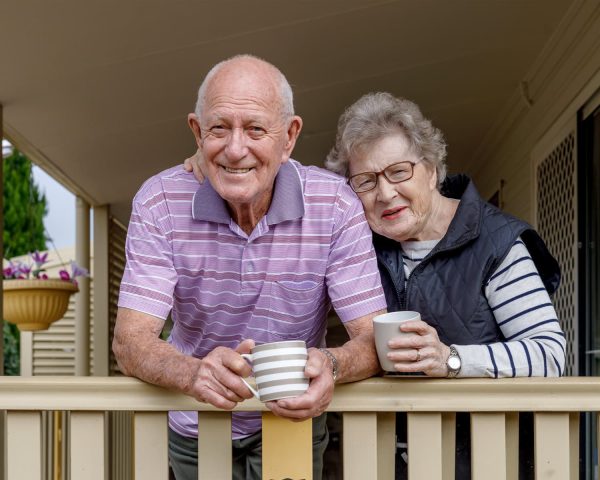 An elderly couple lean on their porch railing holding a cup of tea each. The woman is leaning her head on her husband's shoulder affectionately.