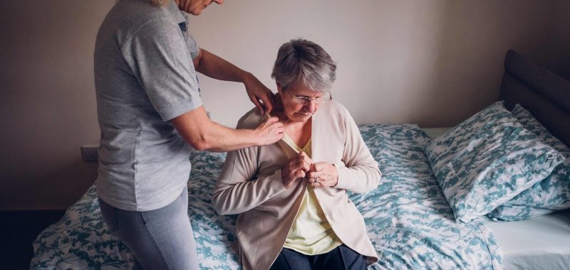 The woman is helping a senior woman dress in her bedroom.