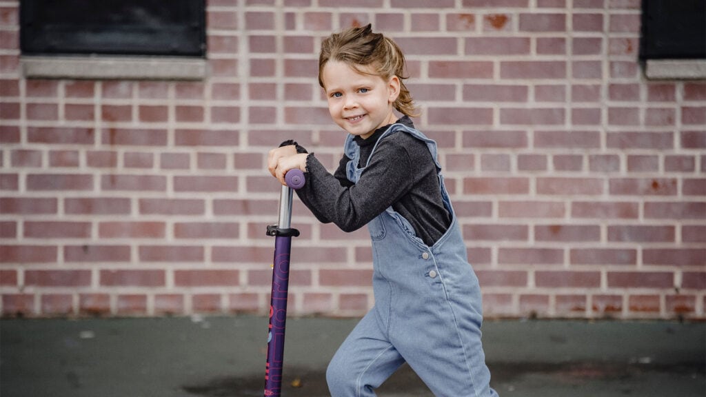 A young blonde child smiles at the camera while riding past on a scooter.